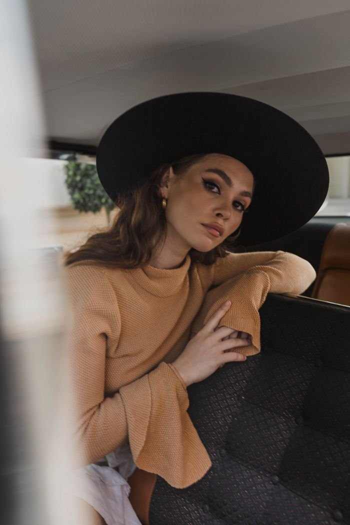 Stylish woman posing in car interior wearing a black hat and beige sweater.